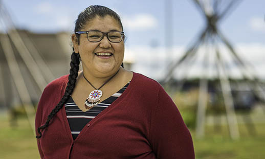 A woman in glasses smiles while looking off to the right of the image. She is wearing a maroon, button up sweater, a black and white striped t-shirt and a beaded necklace. He hair is in a braid. In the background, two teepee frames can be seen. 