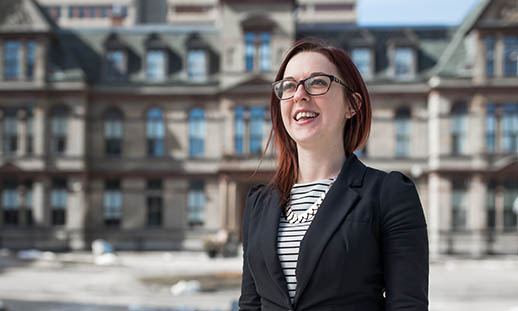 A young woman stands in front of a large, sandstone, townhall building. She is smiling and looking to the top, left of the image. She is wearing a black blazer and a black and white striped t-shirt. Larger buildings can be seen behind the townhall.