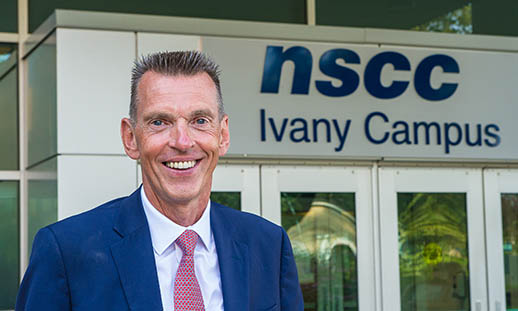 A man, Ray Ivany, stands in front of the doors to NSCC Ivany Campus. He is wearing a blue suit jacket, white shirt and pink tie. He is smiling.
