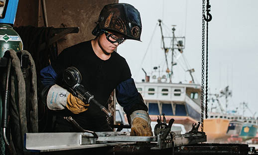 A young man welds a component of a ship. He is wearing a welding helmet (which is open), safety glasses, long leather gloves and a black shirt. In the background, several ships can be seen on land.