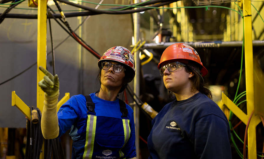 Women Unlimited grad Krista Langille stands next to Megan Miller - whom she mentored - at an Irving shipbuilding facility. They wear safety glasses and hard hats. Krista points to the distance with her right hand.