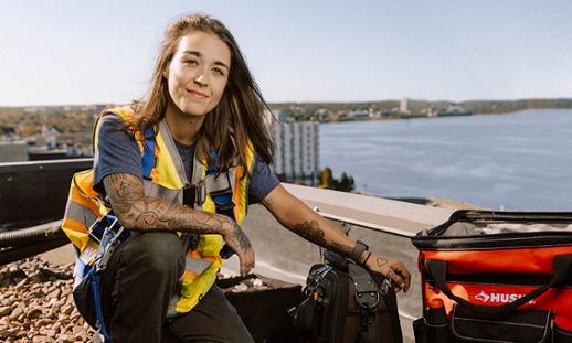 A person wearing a t-shirt, jeans and reflective vest kneels on a rooftop next to a collection of tools. Her left hand rests on a tool bag while she looks at the camera and smiles.