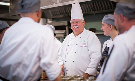 An man wearing a chef's jacket and tall chef's hat stands in a commercial kitchen. Five chefs in pill-box caps and white chef’s jackets are gathered around the other man and appear to be listening to something he is saying. Jars can be seen on the workspace in front of them.