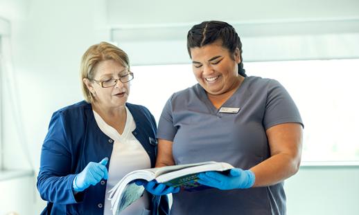 A student wearing nursing scrubs holds an open textbook and looks at it while smiling. An instructor stands next to her and looks at the textbook with her.