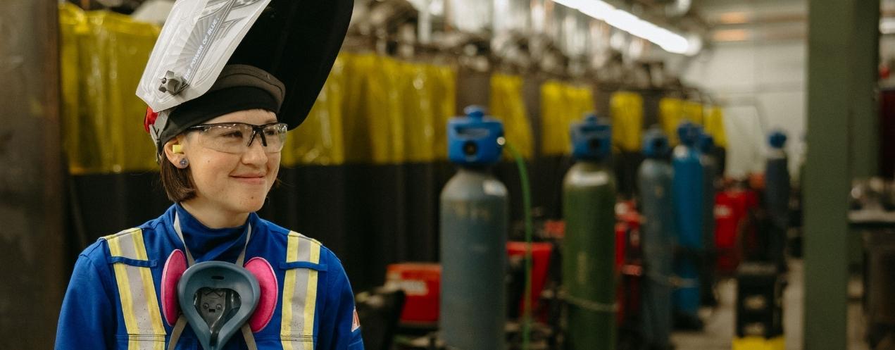 A person wearing personal protective equipment smiling, with welding training equipment visible in the background.