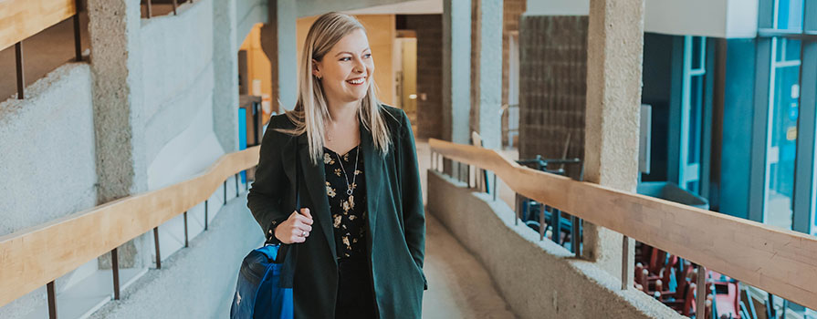Chrissy is an NSCC grad and student award recipient. She is pictured walking up a ramp inside a campus. She is looking toward the window on the right and is smiling.