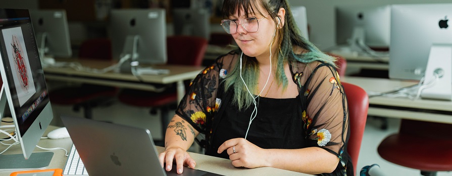 A graphic design student with long hair and glasses sits at a desk and uses a laptop.