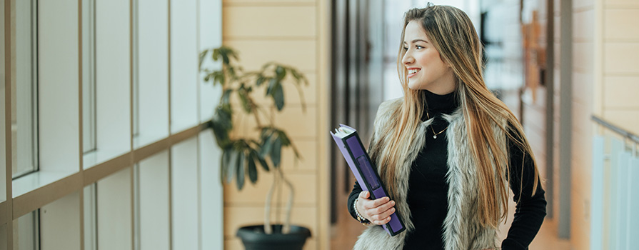 A student with long blond hair walks down an NSCC hallway; she's holding books and smiling.