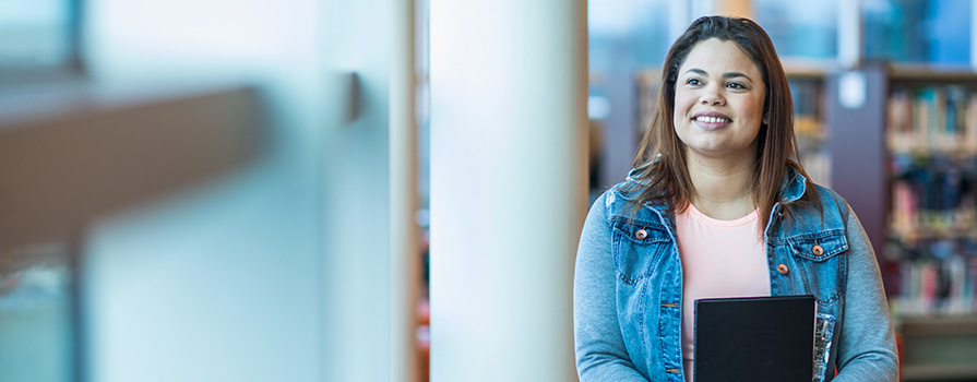 A female student is sitting in an NSCC library; she's looking on and smiling.