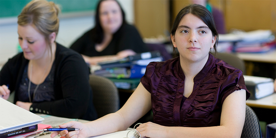 Close up image of two female students at their desk paying attention and taking notes during class with other students also taking notes in the background.