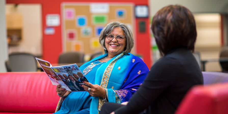 Two women sit on a red couch together smiling and talking.