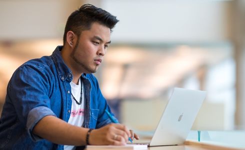A student uses a laptop and takes notes using a pen and paper.