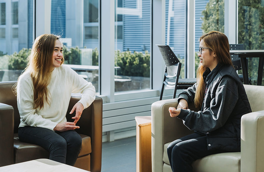 Two students chatting in chairs in an atrium.