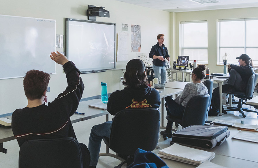 Picture of a teacher at the front of a class of students with one student raising a hand.