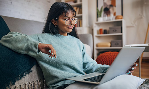 A wearing glasses and a sweater sits comfortably on a couch and looks at a laptop in her living room.
