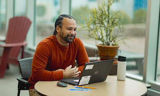 A student wearing a sweater sits at a table in a well-lit space and looks at a laptop.