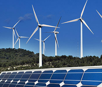 Seven wind turbines against a clear sky tower over a forest, with solar panels pictured in the foreground.