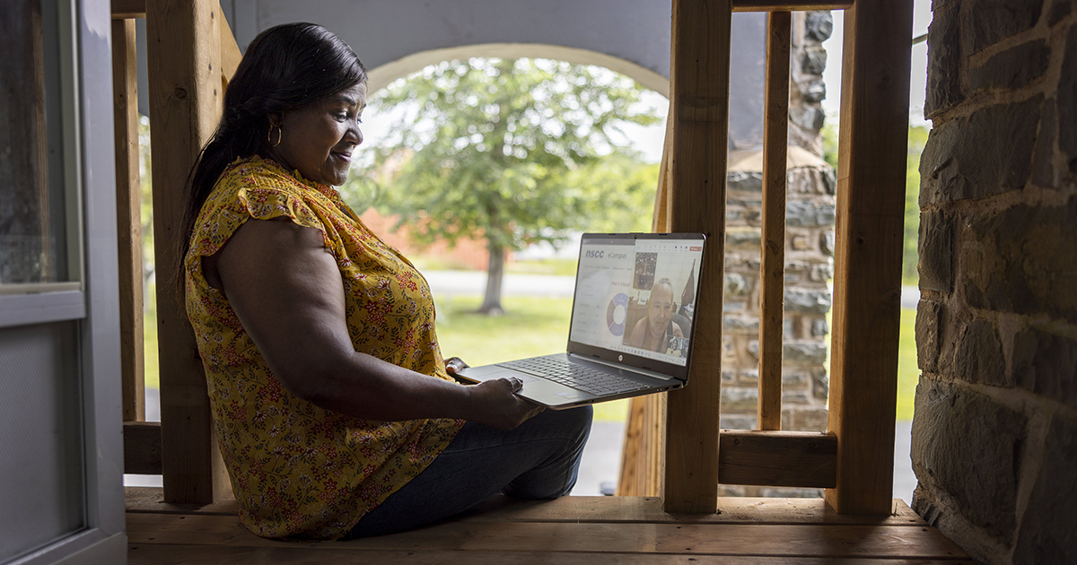 NSCC grad Donna Johnson wears a blouse and jeans and sits on porch stairs while taking part in an online meeting on her open laptop.