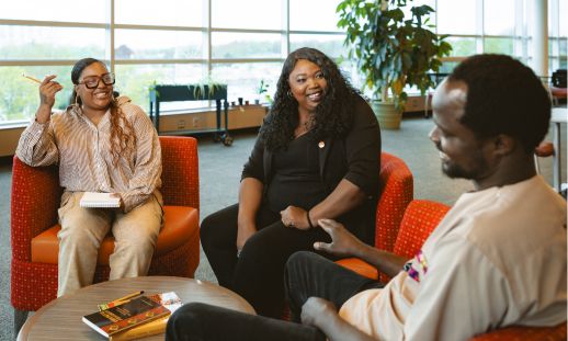 Three people sitting in a library with notepads and books. 