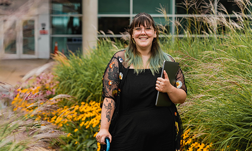 A student stands in front of NSCC Ivany Campus's main entrance and smiles while holding a laptop under her left arm.