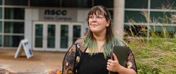 A woman wearing glasses and a black top holds a closed laptop under her left arm and gazes to her right while standing in front of Ivany Campus.