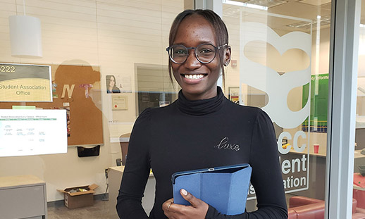 A student wearing a turtleneck and glasses smiles while holding an iPad and standing in front of the Student Association Office at Ivany Campus.