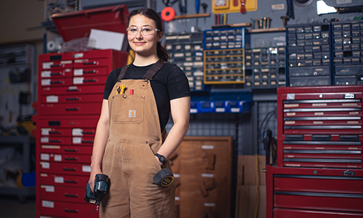 Hilary Ward stands in a construction workshop with a slight smile while wearing a t-shirt, overalls and protective eye wear and carrying a drill.