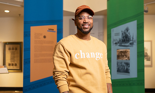 NSCC graduate Paul Adams stands in front of a series of colourful banners indoors at the Black Cultural Centre for Nova Scotia. He smiles at the camera and wears a ballcap, glasses and a sweater with the word change across the front in bold letters.