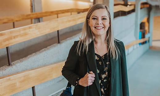A college student stands indoors on a long ramp at an NSCC campus. She wears a blazer and carries a laptop bag over her right shoulder while smiling at the camera.