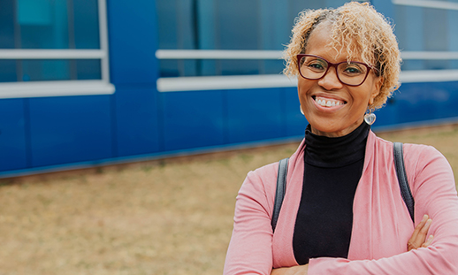 A student wearing a turtleneck, cardigan, earrings, glasses and a backpack stands in front of an NSCC campus with her arms crossed and smiles at the camera.