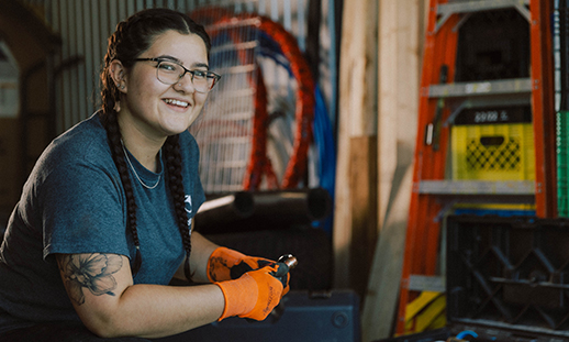 NSCC grad Taylor O'Hearn wears a t-shirt, jeans, glasses and safety gloves. She kneels in a workshop setting and holds a tool while smiling at the camera.