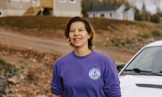 NSCC grad April Googoo stands outdoors in a sweatshirt and gazes ahead while smiling. Behind her are homes and a parked car.