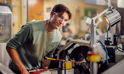 NSCC grad Bryden stands at a machine in a machine shop at an NSCC campus. He wears a long-sleeved sweatshirt and safety goggles and smiles at the camera.