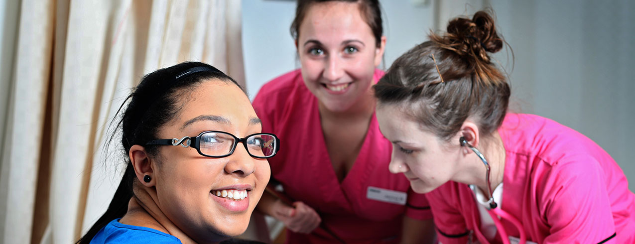 Three Continuing Care students dressed in scrubs practice taking one another’s blood pressure while smiling at the camera.
