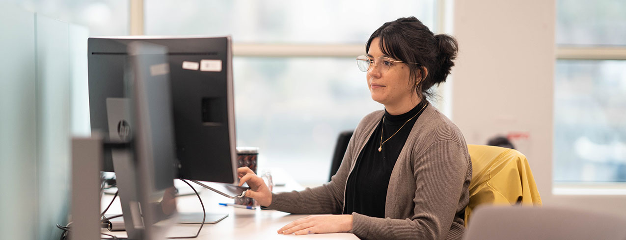 A Digital Marketing professional woman wearing glasses sits at her desk working on her computer. 