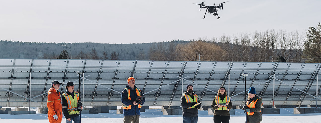 A group of students in the Geomatics Engineering Technology program are seen outside in front of solar panels, wearing safety vests using drones to capture data.