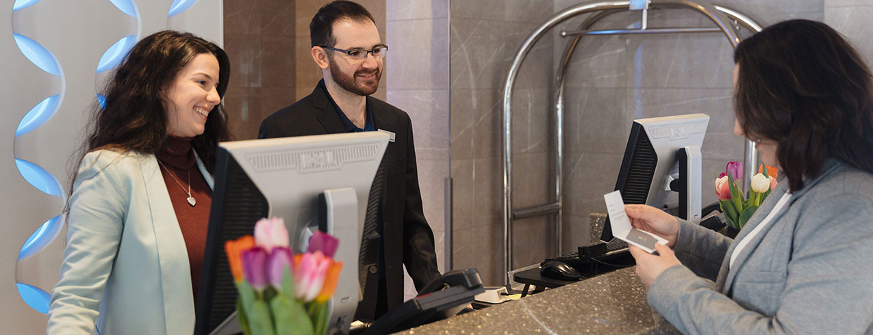 2 hotel and restaurant management students help a customer at the front desk of a hotel.