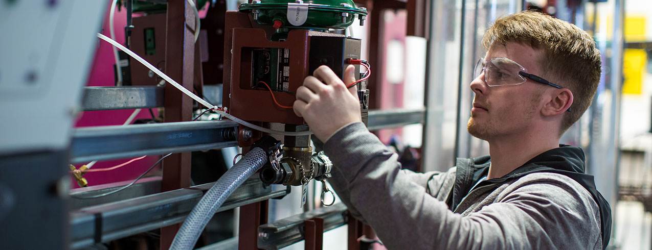 An Instrumentation Technician student is seen working on an electrical panel wearing safety goggles in a factory-type setting. 