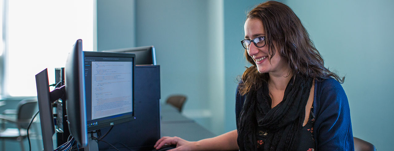 A student in IT Programming with long brown hair and glasses smiles at her computer while she works. 