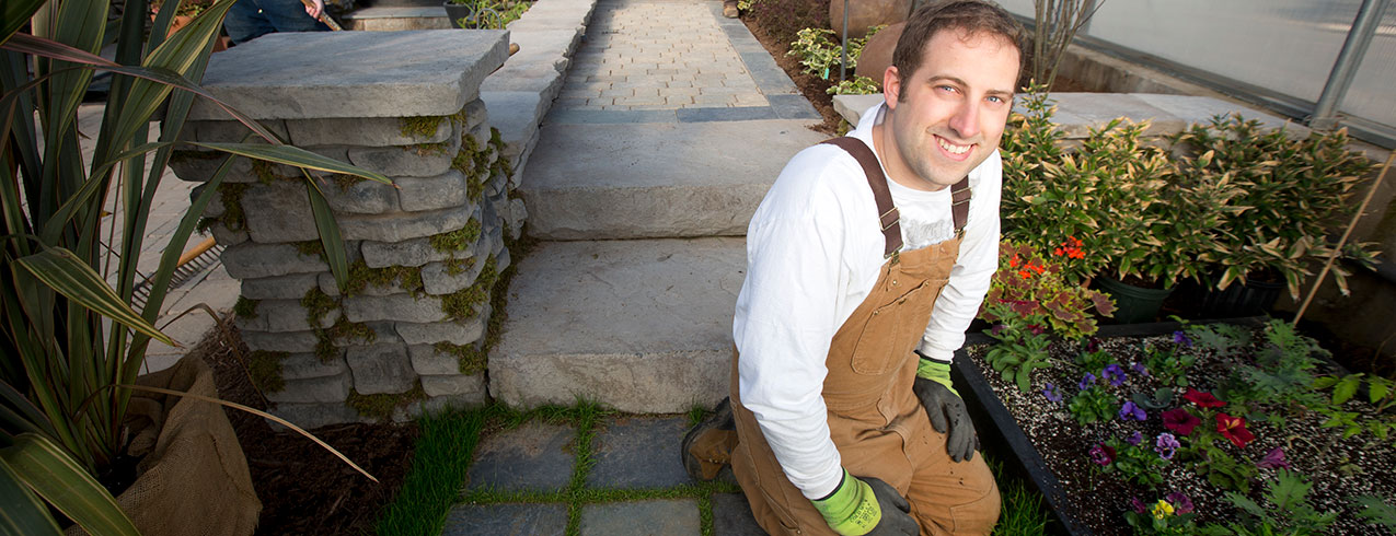 A Landscape Technician student in gardening gloves, wearing brown overalls is seen working on a garden while smiling up at the camera.