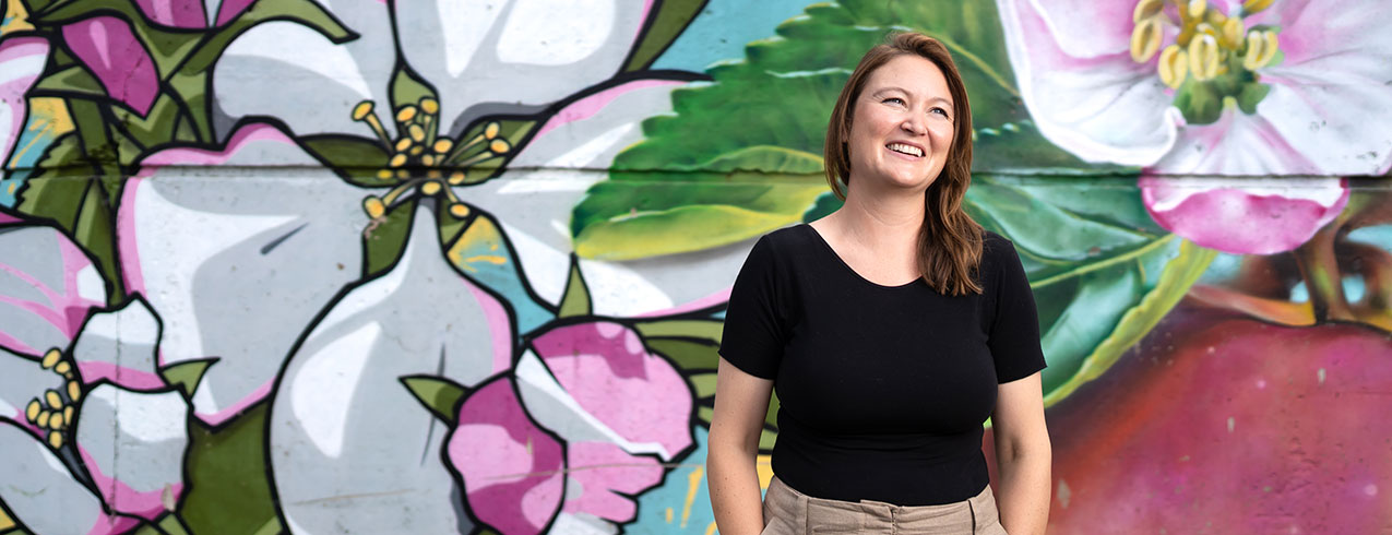 A Nonprofit Leadership student stands in front of a colourful mural painting of flowers and is seen with her hands in her pockets smiling up at the camera.