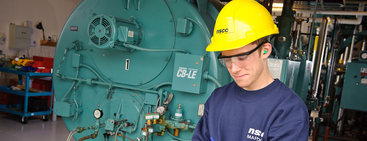 A Power Engineering Technology student in a bright yellow hard hat stands next to a massive machine and looks down at his clipboard.
