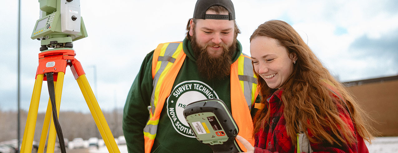 Two smiling Survey Technician students stand outside next to their survey machine looking down at the reading on their connected tablet device. 