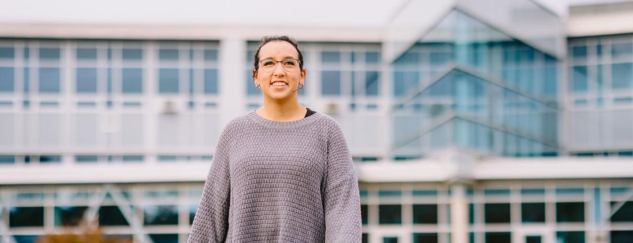 A student stands in front of an NSCC campus - facing away from the campus - and smiles.
