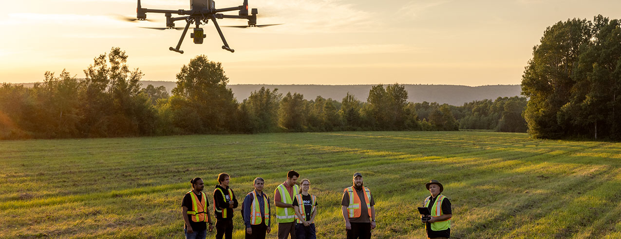 A group of students stand around in a field looking up while one student operates a drone seen above them.