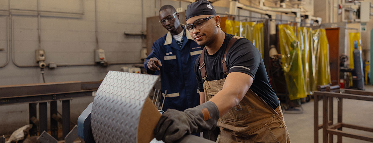 A Metal Fabrication student in work overalls, gloves and safety gear is seen working on a machine while his faculty watches what he's doing. 
