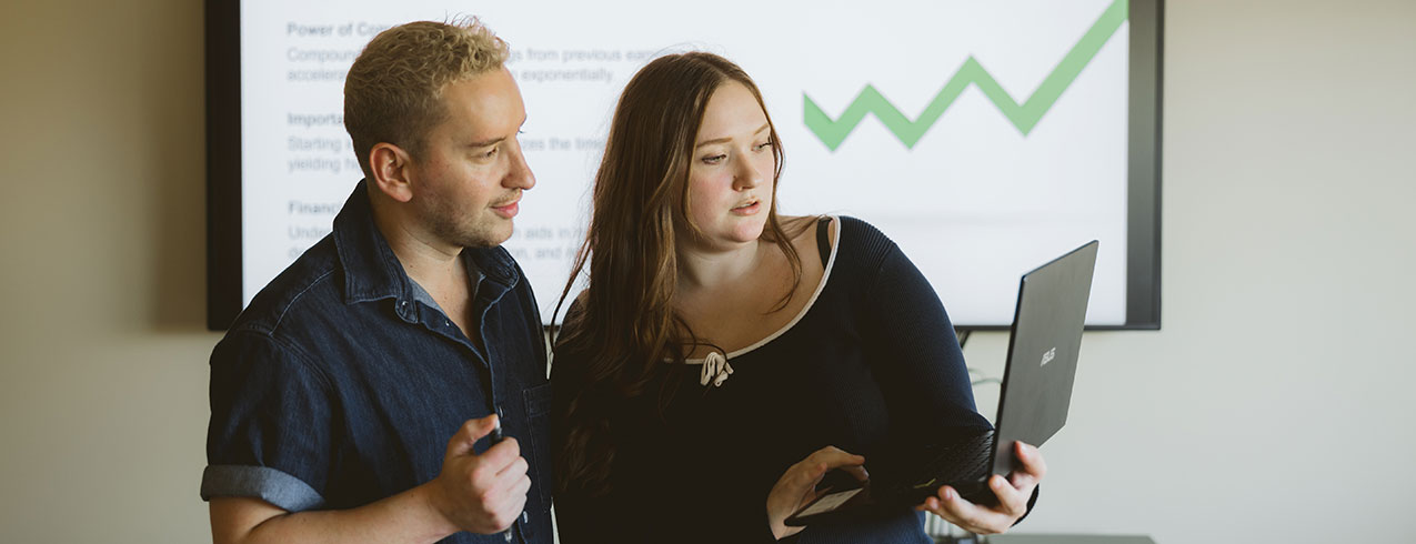 Two NSCC Business Administration students stand together and look at a laptop screen while its being projected on the wall behind them.