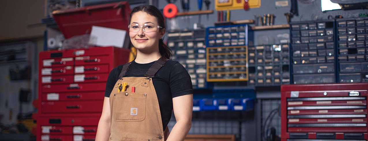 A student wearing overalls and safety glasses stands in a workshop in front of a wall of tools and tool storage.