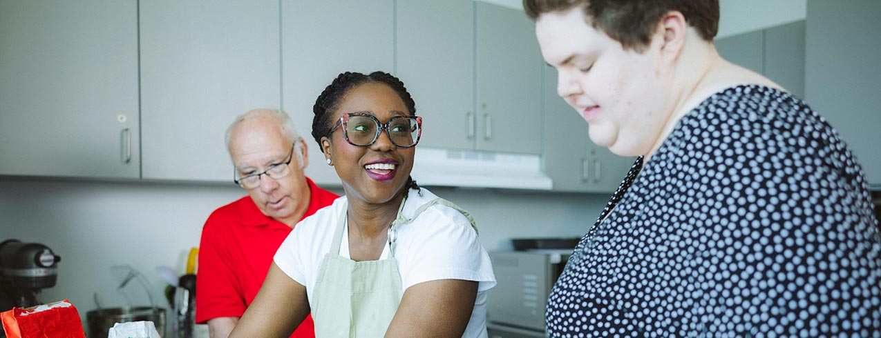 A disability support professional helps two clients bake a batch of cookies in a kitchen.