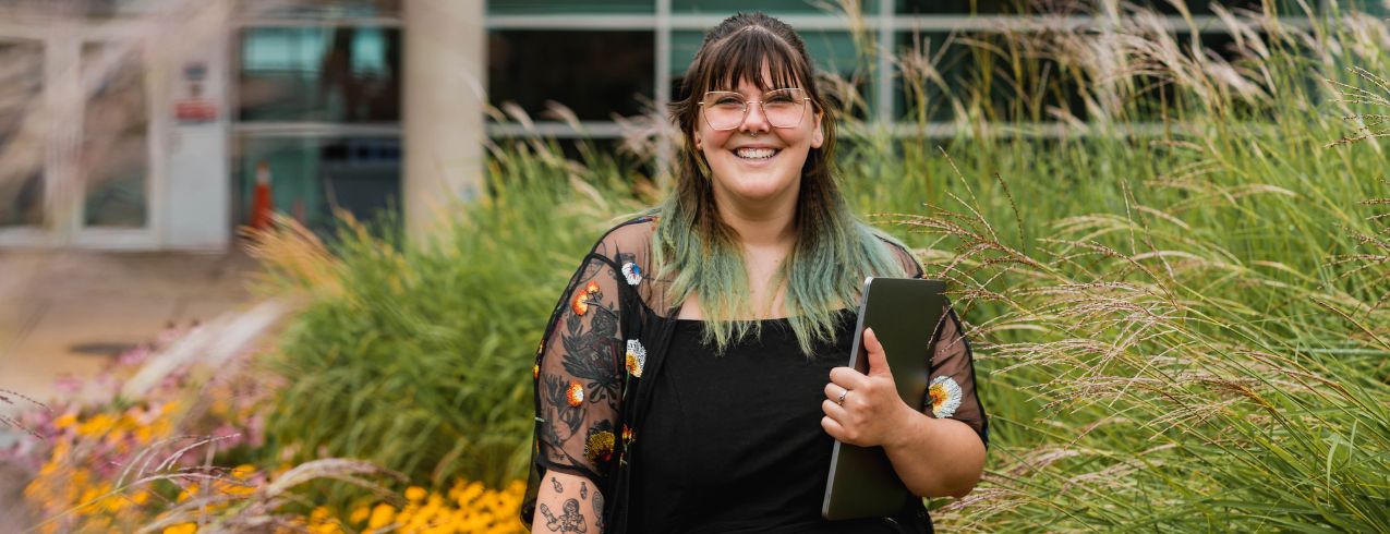 Image of a person walking outside of an NSCC campus, smiling, holding a laptop in their hands. 